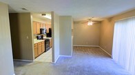 This single-floor space is thoughtfully laid out, offering a clear distinction between the living and kitchen areas. The living room features wall-to-wall carpeting in a neutral tone, creating a comfortable and cohesive feel. A ceiling fan with an integrated light fixture serves as a practical centerpiece overhead, providing both airflow and ambient lighting. Neutral-toned walls throughout the space contribute to a clean, understated aesthetic that suits a variety of décor styles.

The kitchen is accessible via an open passageway and transitions to tile flooring, offering a durable and easy-to-clean surface. It is equipped with black appliances, including a range and overhead microwave, alongside warm-toned wooden cabinetry that lines the walls. Generous counter space and ample storage make the kitchen functional for everyday use. Fluorescent overhead lighting ensures the kitchen is well-lit. A partial wall divides the two areas, maintaining a sense of openness while providing subtle separation between the cooking and living spaces. The overall layout is practical and efficient, making good use of the available floor space.