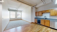 Located at 1004 W Baltimore in the heart of Kansas City, MO, this one-bedroom, one-bathroom unit showcases a distinctive urban loft aesthetic throughout its single-floor layout. The open-concept living and kitchen area greets you with warm hardwood flooring, soaring ceilings with exposed ductwork, and industrial-style ceiling fans that underscore the building's converted character. The kitchen features warm wood cabinetry, dark countertops, a stainless sink, and a built-in beverage cooler, all flowing seamlessly into the spacious living area. Natural light streams in through large, tall windows that frame views of the surrounding Kansas City cityscape, adding brightness and an airy feel to the space. The generously sized bedroom is finished with plush carpeting, a ceiling fan, and its own large windows that continue the theme of natural light and urban views. A dedicated closet off the bedroom provides ample storage. The bathroom offers a clean, functional design with wood-toned cabinetry, a full tub-shower combination, and hardwood-style flooring that ties together the overall warmth of the unit. Bold red baseboards throughout add a subtle but striking design touch, completing this well-appointed urban retreat.