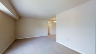 This single-floor residence offers a clean, functional layout designed with simplicity and comfort in mind. The main living area is generously proportioned, featuring wall-to-wall carpeting in a warm neutral tone that lends a cozy, understated feel to the space. Smooth, white walls and a bright ceiling create a sense of openness and airiness throughout the room. A floor-mounted baseboard heating vent is visible along the wall, providing efficient climate control integrated seamlessly into the space. The room flows naturally into an adjoining area, visible through an open passageway, where a classic chandelier with candle-style lights serves as an elegant focal point. This transitional space appears to lead further into the residence, suggesting a well-connected and easy-to-navigate floor plan. Electrical outlets are conveniently positioned throughout the walls, ensuring practical usability of each area. The overall ambiance is bright, neutral, and move-in ready, offering a versatile blank canvas well-suited to a variety of interior styles and furnishing preferences. The consistent use of light tones across the floors, walls, and ceilings contributes to a cohesive, welcoming atmosphere throughout the entire level.