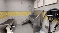 This single-floor pet grooming space is thoughtfully designed to accommodate both pets and their handlers with ease and efficiency. The centerpiece of the room is a large, professional-grade stainless steel dog bathing tub, complete with a built-in walk-up ramp that allows pets to access the tub comfortably without being lifted. A flexible handheld sprayer hose is mounted directly above the tub, enabling thorough and precise rinsing. The walls are finished in neutral gray subway tiles accented by a decorative band of yellow-green mosaic tiles, adding a touch of color and visual interest to the utilitarian space. The flooring is a smooth, easy-to-clean gray surface well-suited for a wet environment. Charming pet-themed artwork adorns the upper walls, lending a welcoming and friendly atmosphere. Additional grooming equipment, including what appears to be a grooming table, is visible along the perimeter, suggesting a fully functional workspace. A wall-mounted fire safety device ensures code compliance. Overall, the space balances practicality with a clean, modern aesthetic, making it a well-equipped environment for professional or self-service pet care.