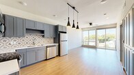 This well-designed one-bedroom, one-bathroom unit offers a thoughtfully arranged open-concept floor plan that maximizes every square foot of its compact footprint. The kitchen is a standout feature, showcasing sleek gray shaker cabinetry, a decorative patterned tile backsplash, white quartz countertops, and a full suite of stainless steel appliances including a refrigerator, electric range, microwave, and dishwasher. Striking black pendant lights hang overhead, adding a modern industrial touch. The kitchen flows seamlessly into the living area, which is bathed in natural light from expansive floor-to-ceiling sliding glass doors that open directly onto a private balcony, offering open views of the surrounding neighborhood and mature trees. Light-toned hardwood-style flooring runs throughout the main living spaces, contributing to the bright and airy ambiance. A ceiling fan overhead adds both function and comfort. The bedroom is a quiet retreat featuring plush carpeting, a ceiling fan, and a dedicated window that allows additional natural light. The bathroom is finished with coordinating blue-gray cabinetry, a white countertop vanity, and wood-style flooring, with convenient direct access to a well-organized walk-in closet with built-in shelving.