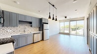 This well-designed one-bedroom, one-bathroom unit offers a thoughtfully arranged open-concept floor plan that maximizes every square foot of its compact footprint. The kitchen is a standout feature, showcasing sleek gray shaker cabinetry, a decorative patterned tile backsplash, white quartz countertops, and a full suite of stainless steel appliances including a refrigerator, electric range, microwave, and dishwasher. Striking black pendant lights hang overhead, adding a modern industrial touch. The kitchen flows seamlessly into the living area, which is bathed in natural light from expansive floor-to-ceiling sliding glass doors that open directly onto a private balcony, offering open views of the surrounding neighborhood and mature trees. Light-toned hardwood-style flooring runs throughout the main living spaces, contributing to the bright and airy ambiance. A ceiling fan overhead adds both function and comfort. The bedroom is a quiet retreat featuring plush carpeting, a ceiling fan, and a dedicated window that allows additional natural light. The bathroom is finished with coordinating blue-gray cabinetry, a white countertop vanity, and wood-style flooring, with convenient direct access to a well-organized walk-in closet with built-in shelving.