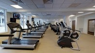 Located at 515 West Ave in Norwalk, Connecticut, this well-appointed fitness center offers a comprehensive workout environment on a single, open floor. The facility is designed with functionality and accessibility in mind, featuring a bright, airy interior illuminated by a combination of large windows that invite natural light and a recessed drop-ceiling lighting grid that ensures consistent illumination throughout the space. The main exercise floor is expansive, with rows of modern cardio machines — including treadmills, upright bikes, and elliptical trainers — arranged to maximize both capacity and ease of movement. Equipment from leading brands such as Life Fitness is prominently featured, reflecting a commitment to quality. The neutral-toned flooring and clean wall finishes contribute to a professional and motivating atmosphere. Mounted monitors are positioned throughout the space, allowing members to stay entertained or informed during their workouts. The layout flows intuitively, with clear sightlines across the floor that make navigation easy. Mirrors along the walls further enhance the sense of openness. Overall, the space presents a polished, well-maintained environment suited for a wide range of fitness routines.