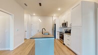 Located at 4665 Whitfield Ave in Sarasota, Florida, this single-story home presents a clean, contemporary design throughout. The open-concept kitchen serves as the centerpiece of the living area, showcasing crisp white shaker cabinetry, stainless steel appliances including a side-by-side refrigerator, electric range, and built-in microwave. A large central island with a quartz countertop and matte black undermount sink provides generous prep and seating space. A geometric white tile backsplash adds a modern touch, while light wood-look flooring flows throughout the main living areas. The bedrooms feature plush carpeting, neutral wall tones, and tall windows fitted with horizontal blinds that allow ample natural light. Walk-in closet space is outfitted with wire shelving systems for organized storage. The bathroom is appointed with large-format white wall tile, a glass-enclosed walk-in shower, and matte black fixtures that complement the home's modern aesthetic. A dedicated laundry room includes a full-size washer and dryer. Recessed lighting and pendant fixtures provide consistent illumination throughout, contributing to the home's bright and welcoming atmosphere.
