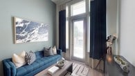 This single-floor space showcases a thoughtfully designed living area that immediately draws attention with its notably tall ceilings, which amplify the sense of openness and volume. Floor-to-ceiling windows and a glass-paneled door flood the room with generous natural light, while navy blue drapes with cream headers frame the windows elegantly, offering both style and light control through adjustable horizontal blinds.

The walls are painted in a soft sage green, creating a calming, cool-toned atmosphere that is complemented by the rich teal tufted sofa positioned as the focal point of the seating area. Geometric patterned throw pillows in black, white, and cream add visual interest against the sofa, while a textured Moroccan-style area rug grounds the space beneath a sleek, low-profile coffee table.

A large abstract landscape artwork in muted blue and grey tones adorns the wall, reinforcing the cohesive color story throughout the room. Light wood-toned flooring runs throughout, adding warmth and contrast to the cooler hues. The overall ambiance is modern, curated, and serene, balancing contemporary design with a comfortable, livable aesthetic.