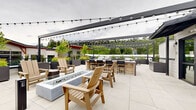 Located in Woodinville, WA, this rooftop terrace is a thoughtfully designed outdoor amenity space offering a wide range of gathering areas and leisure features. The terrace is finished with large-format porcelain tile pavers that lend a clean, contemporary aesthetic throughout the entire level.

Multiple distinct zones are arranged across the rooftop. A lounge area centers around a modern rectangular fire pit, surrounded by warm-toned wood Adirondack-style chairs — an inviting setting for relaxed conversation. Overhead, sleek dark-framed pergolas strung with globe lights provide both structure and ambiance, creating an intimate atmosphere even in an open-air environment.

An outdoor kitchen and bar area offers counter seating with barstool accommodations, ideal for entertaining. Adjacent dining and grilling stations further expand the space's functionality for hosting events of varying scales. Generously sized contemporary planters throughout add lush greenery and natural texture, softening the modern hardscape design.

The perimeter of the terrace features low railing, opening up broad, unobstructed views of the Pacific Northwest treeline and surrounding Woodinville skyline. The combination of practical amenities, thoughtful landscaping, and panoramic natural vistas makes this rooftop terrace a standout communal space designed for year-round enjoyment.