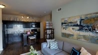 This single-floor residence offers a thoughtfully designed open-concept layout that integrates the living and kitchen areas into one cohesive space. The kitchen is outfitted with dark cabinetry contrasted by a classic white subway tile backsplash, and includes modern black appliances such as a built-in microwave and range. A raised breakfast bar with bar seating provides a casual dining option and serves as a natural dividing element between the kitchen and living areas. Track lighting illuminates the kitchen, adding a warm, functional glow. The living area is anchored by a comfortable gray sectional sofa accented with decorative throw pillows in teal and neutral tones. A large abstract canvas painting adorns the sage-green accent wall, lending an artistic focal point to the room. Rich dark hardwood flooring runs throughout, adding warmth and visual continuity across the space. A wood-framed open shelving unit provides both storage and display space, bridging the two areas stylistically. The overall ambiance is modern and inviting, with a neutral color palette punctuated by carefully chosen accents that create a balanced, livable environment.