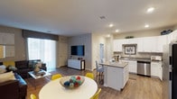 This single-story space showcases an open-concept layout that integrates the dining area, kitchen, and living room into one cohesive, flowing environment. Wood-style flooring runs throughout, lending warmth and visual continuity across all areas.

The dining space accommodates a round white table paired with vibrant yellow chairs, positioned near large sliding glass doors fitted with vertical blinds that allow generous natural light to fill the room. This creates a bright, airy atmosphere throughout the shared living areas.

The kitchen is a standout feature, equipped with white cabinetry, granite or stone-look countertops, and a central island with seating for two. The island provides additional prep space and casual dining flexibility. White appliances and a stainless steel sink complement the clean, modern aesthetic, while recessed lighting ensures ample illumination.

The adjacent living area offers a dedicated media wall and additional storage via a white credenza. Recessed lighting continues throughout, maintaining a consistent, well-lit environment. Neutral gray walls provide a versatile backdrop that complements the bright cabinetry and natural wood tones, creating a balanced and functional living space.