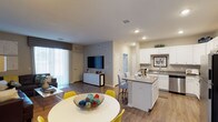 This single-story space showcases an open-concept layout that integrates the dining area, kitchen, and living room into one cohesive, flowing environment. Wood-style flooring runs throughout, lending warmth and visual continuity across all areas.

The dining space accommodates a round white table paired with vibrant yellow chairs, positioned near large sliding glass doors fitted with vertical blinds that allow generous natural light to fill the room. This creates a bright, airy atmosphere throughout the shared living areas.

The kitchen is a standout feature, equipped with white cabinetry, granite or stone-look countertops, and a central island with seating for two. The island provides additional prep space and casual dining flexibility. White appliances and a stainless steel sink complement the clean, modern aesthetic, while recessed lighting ensures ample illumination.

The adjacent living area offers a dedicated media wall and additional storage via a white credenza. Recessed lighting continues throughout, maintaining a consistent, well-lit environment. Neutral gray walls provide a versatile backdrop that complements the bright cabinetry and natural wood tones, creating a balanced and functional living space.