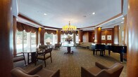 This sophisticated single-floor space presents a refined, hotel-style lounge and lobby area designed with classic elegance and attention to architectural detail. The interior is defined by rich, warm wood paneling that lines the walls and wraps around prominent cylindrical columns, creating a cohesive and stately atmosphere throughout. A striking chandelier with amber-toned shades serves as the centerpiece of the main lounge area, suspended above a round pedestal table adorned with a floral arrangement, anchoring the space with understated grandeur. The curved layout of the main lounge is accentuated by ornate, draped valances framing the expansive floor-to-ceiling windows and sliding glass doors, which flood the interior with natural light and offer unobstructed views of the outdoor patio, complete with seating and landscaped surroundings. A glossy black baby grand piano adds a cultural and artistic focal point to one side of the lounge. Multiple seating arrangements — including upholstered armchairs, sofas, and intimate table-and-chair groupings — are thoughtfully distributed to encourage both social gathering and quiet retreat. The corridors feature polished stone flooring with decorative inlays, transitioning to patterned carpeting in the seating areas. Recessed lighting and table lamps provide warm, layered illumination throughout, contributing to an inviting and refined ambiance.