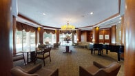This sophisticated single-floor space presents a refined, hotel-style lounge and lobby area designed with classic elegance and attention to architectural detail. The interior is defined by rich, warm wood paneling that lines the walls and wraps around prominent cylindrical columns, creating a cohesive and stately atmosphere throughout. A striking chandelier with amber-toned shades serves as the centerpiece of the main lounge area, suspended above a round pedestal table adorned with a floral arrangement, anchoring the space with understated grandeur. The curved layout of the main lounge is accentuated by ornate, draped valances framing the expansive floor-to-ceiling windows and sliding glass doors, which flood the interior with natural light and offer unobstructed views of the outdoor patio, complete with seating and landscaped surroundings. A glossy black baby grand piano adds a cultural and artistic focal point to one side of the lounge. Multiple seating arrangements — including upholstered armchairs, sofas, and intimate table-and-chair groupings — are thoughtfully distributed to encourage both social gathering and quiet retreat. The corridors feature polished stone flooring with decorative inlays, transitioning to patterned carpeting in the seating areas. Recessed lighting and table lamps provide warm, layered illumination throughout, contributing to an inviting and refined ambiance.