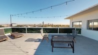 This property features a generous rooftop deck that serves as a standout outdoor living space, offering sweeping panoramic views of a city skyline framed by a clear blue sky and a canopy of mature trees. The deck is finished with light-painted wood planking that gives the space a clean, casual aesthetic, while a white-railed perimeter lined with lush greenery provides both privacy and a polished visual border.

The layout of the rooftop deck accommodates multiple seating arrangements, including a dining table paired with woven chairs and a bench-style seating set, creating distinct zones for outdoor dining and conversation. A lounge chair positioned to take full advantage of the skyline view further enhances the deck's versatility as a relaxation destination. Overhead, a strand of Edison-style string lights spans the width of the deck, adding a warm, ambient glow ideal for evening gatherings.

The open-sky exposure ensures abundant natural light throughout the day, and the elevated position provides an unobstructed vantage point over the surrounding treetops and urban landscape. This rooftop deck functions as a compelling outdoor extension of the living space, well-suited for both casual relaxation and social entertaining.