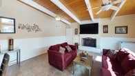 This recreation room is a well-appointed communal gathering space designed for comfort and social interaction. The lounge area serves as the heart of the room, anchored by a classic white-painted brick fireplace with a mounted flat-screen television above it. Plush burgundy sofas and a glass-top coffee table create an inviting seating arrangement, perfect for relaxing or socializing. The vaulted wood-planked ceiling with exposed white-painted beams adds warmth and architectural character to the space, while ceiling fans ensure comfortable airflow throughout.

Natural light pours in generously through large sliding glass doors that frame views of and provide direct access to an outdoor pool area with patio seating, seamlessly connecting the indoor and outdoor environments. The upper transom windows further enhance the bright, airy atmosphere of the room.

A functional kitchenette is tucked along one wall, equipped with a full-size stainless steel refrigerator, microwave, sink, and a light-toned countertop bar with seating for casual dining. A dining area with a glass table and dark wood chairs completes the space. Restroom facilities are also conveniently accessible from within the room. The combination of warm wood tones, neutral walls with wainscoting, and tile flooring gives the space a clean, comfortable, and versatile character.