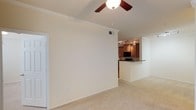 This single-floor residence showcases an open-concept layout that seamlessly connects the living area to the kitchen. The living space features vaulted ceilings adorned with elegant crown molding, adding a refined architectural detail to the room. A ceiling fan with an integrated light fixture is centrally mounted, providing both air circulation and ambient lighting. The neutral, light-toned carpeting runs throughout, contributing to a clean and cohesive aesthetic.

The kitchen is visible through a pass-through opening, defined by a raised dark-topped breakfast bar that serves as a natural divider between the two spaces. Warm, medium-toned wood cabinetry lines the kitchen walls, complemented by track lighting overhead that illuminates the work area effectively. Black appliances add a contemporary contrast to the cabinetry. The walls throughout are finished in a soft, neutral tone, enhancing the sense of brightness and openness. Overall, the space offers a practical and comfortable layout with classic design elements that create a welcoming, functional environment.
