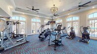 This well-appointed fitness center in Cary, NC is thoughtfully designed to support a wide range of workout routines across two distinct areas. The main exercise room features an elegant tray ceiling with decorative crown molding, ceiling fans, and an ornate chandelier. Large arched windows and glass-paned French doors flood the space with natural light while offering outdoor views, creating an energizing atmosphere. The room is outfitted with a comprehensive selection of cardio equipment, including treadmills, StairMaster climbers, ellipticals, and a stationary bike, all arranged across a bold patterned carpet in deep blue and teal tones. A mirrored wall expands the sense of space and supports strength training with a dedicated dumbbell rack and cable machines nearby. A secondary studio room, finished with light hardwood-style flooring and a bright, neutral color palette, provides a versatile space for group or individual training. It accommodates spin bikes, a rowing machine, kettlebells, stability balls, and other functional fitness accessories. Soft recessed and panel lighting keeps this area well-lit, while arched windows maintain a connection to the outdoors. Together, the two zones create a comprehensive and inviting fitness environment suitable for diverse exercise needs.