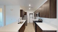 This single-floor space showcases a thoughtfully designed interior with a strong emphasis on modern finishes and functional layout. The kitchen is a standout feature, styled as a galley with generous countertop space finished in crisp white quartz. Rich, dark wood cabinetry lines both sides of the kitchen, extending to the ceiling to maximize storage, and is complemented by sleek brushed hardware. Stainless steel appliances include a gas range with multiple burners, an over-the-range microwave, a dishwasher, and a full-size refrigerator, providing a complete and contemporary cooking environment. The double-basin stainless steel sink is positioned along one counter, offering ample workspace on either side. Light wood-tone flooring runs throughout, adding warmth and continuity to the space. Recessed lighting illuminates the kitchen evenly, enhancing the clean, bright atmosphere created by the white walls and ceilings. A dedicated in-unit laundry area is conveniently accessible directly from the kitchen, housing a stacked washer and dryer unit. Overall, the space presents a cohesive, modern aesthetic that balances practicality with refined design details.