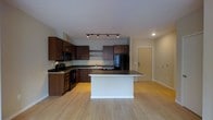 This single-floor space showcases a thoughtfully designed open-concept layout that seamlessly blends functionality with modern aesthetics. The kitchen serves as the centerpiece of the space, featuring rich dark-wood cabinetry that provides generous storage, complemented by dark countertops that add a sleek, contemporary contrast. A central kitchen island with a light-toned surface offers additional workspace and serves as a natural gathering point. Black appliances, including a refrigerator, microwave, range, and dishwasher, lend a cohesive and polished look to the culinary area. Open shelving adds a modern touch while providing accessible display and storage options. Track lighting overhead ensures the kitchen is well-lit, while recessed lighting extends throughout the rest of the space for a clean, uncluttered ceiling aesthetic. Light hardwood flooring runs throughout the unit, adding warmth and continuity across the open floor plan. The space flows naturally from the kitchen into the adjoining living area, creating an expansive, airy feel. Neutral wall tones keep the atmosphere bright and versatile. A solid entry door with keypad access is visible in the background, reinforcing both the security and contemporary character of this well-appointed space.
