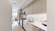This contemporary single-floor residence is thoughtfully designed with a clean, modern aesthetic and an emphasis on natural light. The galley-style kitchen is a standout feature, showcasing floor-to-ceiling cabinetry in a warm wood-grain finish, stainless steel appliances including a gas range, built-in microwave, refrigerator, and dishwasher, complemented by white quartz countertops and a subway tile backsplash. Recessed lighting runs the length of the kitchen, which opens toward a bright living area bathed in natural light from large windows. The bedroom is generously proportioned, featuring dark hardwood flooring, a ceiling fan, and full-height glass doors that open to an outdoor balcony with neighborhood views. A secondary seating area within the bedroom benefits from its own set of large windows, further enhancing the sense of openness. The bathroom offers a clean, minimalist design with light tile flooring, a glass-enclosed shower, and neutral wall tones. A compact dining nook adjacent to a secondary kitchenette provides a versatile, efficient living arrangement. Throughout the residence, white walls, recessed lighting, and an open layout create a bright, airy ambiance that is both functional and inviting.