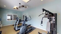 Located at 7457 N Kensington Way in McCordsville, Indiana, this single-floor fitness facility offers a dedicated space designed to support a comprehensive workout routine. The main exercise room is bright and inviting, finished with cool blue-toned walls that create a calm, energizing environment. The space is outfitted with multiple cardio machines, including elliptical trainers and a treadmill, all positioned for easy access and comfortable use. A multi-station strength training machine is mounted along one wall, providing a range of resistance and cable exercises without requiring additional equipment. Overhead, a stylish dual-blade ceiling fan circulates air throughout the room, complementing the recessed lighting and a skylight that draws in natural light from above. The flooring is fitted with protective exercise mats beneath the equipment, ensuring safety and stability during workouts. Motivational wall decor adds a personal touch to the space. Double doors offer access to an adjacent area, adding to the functional flow of the facility. Overall, this fitness space strikes a practical balance between well-selected equipment and a comfortable, welcoming atmosphere suited for daily exercise.
