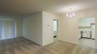 This single-floor space presents a clean and understated interior design with a neutral color palette throughout. The main living area is finished with wall-to-wall carpeting in a soft, muted tone that complements the crisp white walls, creating a calm and cohesive atmosphere. A textured popcorn ceiling adds a classic touch overhead, while an elegant chandelier with candelabra-style lighting serves as a refined focal point, lending a subtle sense of formality to the space. A partial interior wall divides the open area, adding architectural interest and a sense of defined zones without fully enclosing the space. Through a doorway, a secondary room is visible, featuring what appears to be built-in cabinetry or closet doors with a bright, clean finish, suggesting well-organized storage solutions. Electrical outlets and light switch plates are positioned thoughtfully along the walls, and neutral baseboards frame the flooring throughout. The overall ambiance is bright, tidy, and move-in ready, offering a versatile blank canvas well-suited for a variety of interior configurations and personal styles.