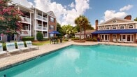 Located at 100 Granby Crossing in Cayce, South Carolina, this outdoor pool area serves as a central amenity space within a well-maintained residential community. The centerpiece is a generously sized swimming pool with clear, blue-green water, bordered by a classic brick paver deck that provides ample space for lounging and socializing. Towering palm trees are thoughtfully positioned throughout the area, lending a resort-like atmosphere and natural shade. The pool is accessible via stainless steel entry rails and features clearly marked depth indicators, with signage noting safety regulations including a no-diving policy. Sun loungers and chaise chairs are arranged along the pool's perimeter, and shaded seating areas with navy blue market umbrellas offer comfortable spots for relaxation. A poolside structure — likely a cabana or amenity building — is positioned adjacent to the water, providing additional convenience for guests. The surrounding landscape is richly planted with flowering crepe myrtles, neatly trimmed shrubs, and mature trees that frame the space with vibrant seasonal color. Multi-story residential buildings with balconies, brick facades, and traditional architectural detailing form a backdrop that reinforces the community's cohesive aesthetic. Open to blue skies and natural sunlight, this pool area delivers an inviting and well-appointed outdoor retreat.