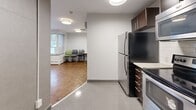Located at 200 Ridley Blvd in Toronto, this single-floor communal space is thoughtfully designed to serve as both a functional kitchen and a shared gathering area. The kitchen is outfitted with dark espresso cabinetry, stainless steel appliances including a full-size refrigerator, electric range, and over-the-range microwave, complemented by a white subway tile backsplash and a sink with under-cabinet storage. The floor transitions from large-format polished grey tiles in the kitchen zone to rich warm-toned parquet hardwood flooring in the lounge and dining area. The main room features grey-painted walls, flush-mount drum ceiling lights, and a wall-mounted flat-screen television. A built-in white counter along one wall provides additional workspace or a serving surface. The space is furnished with folding tables and stackable chairs, offering flexible seating arrangements. A wall-mounted split air conditioning unit ensures year-round climate comfort. Natural light enters through a window overlooking the exterior, and a secured entry door with keypad access provides controlled entry. The overall design is clean, functional, and practical, suited for shared use.