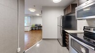 Located at 200 Ridley Blvd in Toronto, this single-floor communal space is thoughtfully designed to serve as both a functional kitchen and a shared gathering area. The kitchen is outfitted with dark espresso cabinetry, stainless steel appliances including a full-size refrigerator, electric range, and over-the-range microwave, complemented by a white subway tile backsplash and a sink with under-cabinet storage. The floor transitions from large-format polished grey tiles in the kitchen zone to rich warm-toned parquet hardwood flooring in the lounge and dining area. The main room features grey-painted walls, flush-mount drum ceiling lights, and a wall-mounted flat-screen television. A built-in white counter along one wall provides additional workspace or a serving surface. The space is furnished with folding tables and stackable chairs, offering flexible seating arrangements. A wall-mounted split air conditioning unit ensures year-round climate comfort. Natural light enters through a window overlooking the exterior, and a secured entry door with keypad access provides controlled entry. The overall design is clean, functional, and practical, suited for shared use.