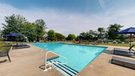 Located at 1300 Fayette St in Conshohocken, Pennsylvania, this outdoor recreational area is centered around a generously sized swimming pool with crystal-clear blue water. The main pool is rectangular in shape and features entry ladders, depth markers along the edges, and a wide concrete surround that provides ample space for lounging. Multiple chaise lounge chairs and patio furniture, including shaded umbrella tables, are arranged around the pool deck, offering comfortable spots for relaxation. A separate baby pool with its own dedicated area is also present, making this space family-friendly. The pool complex includes a single-story clubhouse building with a sliding glass door entry and a charming blue accent door, with a small outdoor table and umbrella positioned along its exterior wall. The grounds are beautifully landscaped with mature evergreen and deciduous trees that provide natural shade and privacy, along with manicured shrubs and well-kept grass areas. A fenced perimeter encloses the pool area for safety, while the surrounding greenery creates a tranquil, park-like atmosphere. Open skies above the space allow for abundant natural light throughout the day, enhancing the inviting outdoor ambiance of this community amenity.