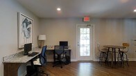 This single-floor space is thoughtfully designed to serve as a practical and comfortable work environment. The interior is finished with warm hardwood flooring throughout, which pairs well with the neutral, light gray walls to create a clean and professional atmosphere.

A built-in granite countertop workstation runs along one wall, providing ample desk space accommodating multiple computer workstations and ergonomic task chairs. The counter's stone surface adds a polished, durable quality to the workspace. Recessed ceiling lighting ensures consistent illumination across the area, while a stylish table lamp with a metallic base adds a warm accent to the workspace.

A glass-paneled exterior door with a classic multi-lite design floods the room with natural light and offers a view of the surrounding outdoor greenery. Positioned near the door is a high-top bistro table with stools, offering an informal spot for casual seating or collaboration.

The space is equipped with an emergency exit sign above the door, reflecting its commercial or community-use nature. Clean white trim and baseboards frame the room neatly, lending a crisp, well-maintained finish throughout. Overall, this space delivers a functional yet welcoming environment well-suited for shared or business use.