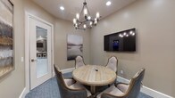 This single-floor meeting space is designed to facilitate intimate gatherings and focused collaboration. The room centers around a round, reclaimed-wood table surrounded by four upholstered armchairs with tufted backs and nailhead trim detailing, offering a refined yet comfortable seating arrangement. Overhead, a wrought-iron chandelier with cylindrical glass shades provides warm, ambient lighting complemented by recessed ceiling fixtures.

A wall-mounted flat-screen display is positioned prominently on one wall, making the space well-suited for presentations or media sharing. The walls are finished in a warm taupe tone, lending a neutral and professional atmosphere, while framed landscape artwork adds visual interest and a sense of calm throughout.

Patterned blue-gray carpet covers the floor, adding texture and a subtle decorative element. Glass-paneled French doors connect the meeting room to an adjacent computer workspace, which is visible through the glazing and features ergonomic seating, desktop workstations, and additional chandelier lighting. This transparent connection maintains visual openness while preserving acoustic separation. The overall design balances functionality with polished, transitional-style aesthetics, creating an environment suited for professional meetings or private consultations.