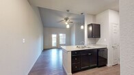 This single-story residence offers 2 bedrooms and 3 bathrooms across a thoughtfully designed floor plan. The open-concept kitchen and living area serves as the heart of the home, highlighted by soaring vaulted ceilings and a ceiling fan that enhances the sense of spaciousness. The kitchen is equipped with dark espresso cabinetry, granite-style countertops, a peninsula with a double sink, and black appliances including a refrigerator and range. An arched architectural detail connects the kitchen to the adjoining areas, adding a decorative touch. Wood-style flooring runs consistently throughout the main living spaces and bedrooms, lending a cohesive and contemporary feel. The living area features a door with an adjacent window that opens to an outdoor balcony, allowing natural light to fill the room. Both bedrooms are comfortably sized, with the primary bedroom offering its own private bathroom featuring dark cabinetry, a granite-style vanity, a tall linen tower, and direct access to an adjoining room. The additional bathrooms include a tub-shower combination. A utility room, hallway, and closet provide practical storage and functional support throughout. Neutral wall tones and clean finishes complete the modern, move-in-ready aesthetic of this residence.