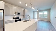 This single-floor residence showcases a thoughtfully designed open-concept layout that seamlessly connects the kitchen and living areas. The kitchen serves as the centerpiece, anchored by a generous island with a speckled white quartz countertop and an undermount sink fitted with a brushed nickel pull-down faucet. A linear pendant light fixture with cylindrical shades illuminates the island, while recessed lighting brightens the surrounding space. Gray shaker-style cabinetry lines the walls, complemented by a white subway tile backsplash and a full suite of stainless steel appliances, including a side-by-side refrigerator, electric range, and over-the-range microwave. Light wood-style flooring extends through the kitchen and into the living area, where large windows and a glazed door invite abundant natural light and outdoor views. A ceiling fan adds comfort to the living space. The bedroom features plush carpet flooring, a ceiling fan, and a well-sized window framing pleasant exterior views. Additional rooms feature light wood-style flooring and bright, neutral-toned walls. The bathroom includes gray vanity cabinetry, a quartz countertop, and a tub-shower combination, maintaining the home's clean, contemporary aesthetic throughout.