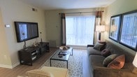 This single-floor space offers a thoughtfully arranged and inviting interior designed for comfortable everyday living. The living room serves as the heart of the space, highlighted by a large picture window dressed with vertical blinds and decorative curtains that flood the room with generous natural light. Hardwood-style flooring runs throughout, adding warmth and a clean aesthetic to the overall design. The living area is well-appointed with a plush sectional sofa, a sleek glass-top coffee table resting on a patterned area rug, and a wall-mounted flat-screen television paired with a coordinating media console. A stylish floor lamp and table lamp provide soft ambient lighting, complementing the natural brightness from the window. Subtle decorative accents, including toss pillows and tasteful tabletop pieces, contribute to the space's polished yet relaxed feel. A wall-mounted HVAC vent ensures year-round climate comfort. The layout is open and efficiently organized, making the most of the available square footage. The overall ambiance is calm and neutral, with a contemporary style that feels both livable and well-curated — ideal for those seeking a functional and aesthetically pleasing environment.