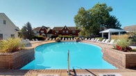 Located at 5303 Wilcox Rd in Dublin, Ohio, this single-floor community amenity space is highlighted by a generously sized outdoor swimming pool that serves as the centerpiece of the property. The pool features a smooth, curved design with crystal-clear blue water and is accessed via a sturdy metal handrail entry point. A wide brick-paved pool deck surrounds the water on all sides, providing ample space for relaxation and recreation. Numerous lounge chairs are arranged along the perimeter, offering residents plenty of options for sunbathing and outdoor leisure. The deck is framed by mature trees that provide natural shade and a lush, green backdrop, enhancing the overall outdoor ambiance. A covered structure is visible at the far edge of the deck, adding a shaded gathering area to the amenity space. The backdrop includes a classic red-brick residential building with traditional peaked rooflines, white-trimmed windows, and well-maintained landscaping featuring ornamental grasses and shrubs. The setting evokes a resort-like atmosphere under open skies, making it an inviting outdoor destination suited for warm-weather enjoyment. The combination of thoughtful design, mature greenery, and well-appointed seating creates a comfortable and attractive communal outdoor environment.