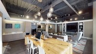 This contemporary single-floor office space blends industrial aesthetics with modern design sensibilities to create a dynamic and functional work environment. The open-plan layout is anchored by a large, live-edge natural wood communal table fitted with multiple computer workstations and white ergonomic seating, making it an ideal hub for collaboration and productivity.

Overhead, an exposed ceiling with visible ductwork, steel beams, and a striking cluster of globe pendant lights lends the space an urban, loft-like character. Track lighting supplements the pendant fixtures, ensuring the workspace is well-lit throughout.

Dark hardwood flooring runs through the main work area, providing a warm contrast to the industrial ceiling elements. Glass partitions divide portions of the floor, allowing natural light to flow through while maintaining visual connectivity across zones.

Toward the rear, a lounge area offers a more relaxed setting, furnished with a sofa, accent seating, and a large wall-mounted display screen. A built-in shelving unit with decorative accessories adds a residential warmth to this social zone. The space also includes a kitchenette or utility area tucked along one side, enhancing day-to-day convenience. Overall, the office strikes a refined balance between professional functionality and inviting design.