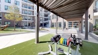 Located in Fort Worth, Texas, this expansive outdoor amenity courtyard is set within a contemporary multi-story residential community on Dakota Street. The space is thoughtfully designed to serve as a central hub for recreation and social activity, seamlessly blending fitness and leisure in an open-air environment.

A standout feature is the dedicated outdoor fitness zone, surfaced with lush artificial turf and equipped with a full complement of strength training equipment, including weight racks, barbells, medicine ball storage, a tire sled, and a multi-functional exercise tower. A sleek metal pergola provides partial shade over the workout area, allowing residents to train comfortably while remaining connected to the outdoors.

Beyond the fitness zone, the courtyard extends into a spacious hardscaped plaza lined with additional pergola structures strung with overhead lighting, shading rows of modern outdoor seating. This area is well-suited for casual gatherings and social events. A dedicated lawn game area, complete with a cornhole board, adds a recreational dimension to the space.

Landscaping throughout the courtyard incorporates native plantings and greenery that soften the clean architectural lines of the surrounding buildings. The multi-story façades feature a mix of white paneling and brick, with private balconies overlooking the courtyard. Abundant natural light fills the space under the open Texas sky, creating a bright and energetic atmosphere year-round.