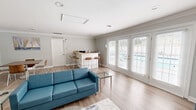 The Kingsbridge Clubhouse in Chesapeake, Virginia offers a well-appointed, single-floor community gathering space designed for comfort and versatility. The interior is finished with light hardwood-style flooring and soft neutral gray walls, accented by crisp crown molding that runs throughout, lending the space a polished, cohesive look.

The main area is thoughtfully arranged into distinct functional zones. A lounge area features teal upholstered seating — including a sofa and armchairs — centered around a glass-top coffee table and an area rug with an abstract pattern. A wall-mounted flat-screen television serves as a focal point in this zone, complemented by decorative circular wall accents. Adjacent to the lounge is a dining area with a warm walnut-toned rectangular table surrounded by mid-century modern wood chairs, ideal for meetings or group gatherings. Nautical-themed artwork adorns the walls, adding a regional coastal character.

A built-in counter with bar-height seating functions as a kitchenette area, providing a convenient space for refreshments. Multiple French doors line the exterior walls, flooding the space with natural light and offering views of an outdoor pool area. Recessed lighting throughout ensures the space remains bright and welcoming. The overall ambiance is clean, modern, and inviting — well-suited for community events, social gatherings, or casual meetings.
