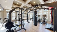 Located at 1050 Old Gilliard Rd in Ridgeville, SC, this fitness center offers a generously sized, single-floor exercise space designed to support a wide variety of workout routines. The room's architecture is immediately striking, featuring elegant coffered ceilings with recessed lighting and contrasting dark insets that lend a polished, upscale aesthetic. Large ceiling fans provide airflow throughout the space, complementing the abundant natural light that streams in through floor-to-ceiling windows lining multiple walls and offering views of the surrounding landscape.

The flooring throughout consists of warm-toned wood-look planks, creating a clean and inviting atmosphere. Full-length mirrors are strategically positioned to enhance the sense of openness and aid users during exercise. The facility is comprehensively outfitted with a diverse selection of equipment, including a row of stationary cycling bikes, treadmills, elliptical machines, and cable weight systems. A functional training rig anchors the central area, complete with a punching bag, monkey bars, and battle rope attachments. Free weights, kettlebells, medicine balls, and adjustable benches round out the strength training offerings. A wall-mounted flat-screen TV adds a modern convenience to the space, making this fitness center a thorough and well-appointed facility.