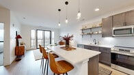 This thoughtfully designed one-bedroom, one-bathroom residence offers a cohesive blend of modern aesthetics and functional living across a single floor. The open-concept kitchen and living area serves as the heart of the unit, anchored by a large quartz-topped island with seating for multiple guests and illuminated by a trio of elegant pendant lights. Dark-toned cabinetry pairs seamlessly with a white tile backsplash, stainless steel appliances, an undermount sink with a matte black faucet, and open floating shelves that add both style and utility. Natural light pours into the living room through floor-to-ceiling black-framed windows and a sliding glass door that opens to a private balcony, offering open outdoor views. The hardwood flooring throughout the main living areas adds warmth and continuity. The bedroom is a serene retreat finished with plush carpeting and direct access to a spacious walk-in closet complete with dual hanging rods. The full bathroom features large-format tile flooring, a dark wood vanity with a white countertop, a wide mirror with modern sconce lighting, and ample storage. A dedicated in-unit laundry space houses a stacked washer and dryer, adding everyday convenience to this well-appointed home.