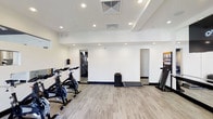 This fitness facility is thoughtfully designed to accommodate a variety of workout routines within a well-organized, single-level layout. The space features light wood-look flooring throughout, providing a clean and durable surface suited to high-traffic exercise use. Recessed lighting is embedded into the stepped ceiling, ensuring the room is evenly and brightly illuminated without harsh shadows.

The main workout area is equipped with a row of stationary cycling bikes positioned along one side, making it well-suited for cycling sessions or spin-style training. A treadmill is also present, offering cardio options for users. Weight plates are neatly stacked in the center of the room, indicating available free-weight training resources. The white walls are largely unobstructed, contributing to a spacious and open atmosphere.

Two doorways lead to additional adjoining areas — one appearing to connect to a mirrored weight room or strength training zone, and another opening to a separate room. The overall ambiance is clean, minimal, and functional, reflecting a contemporary design approach focused on providing an efficient and comfortable environment for physical fitness.