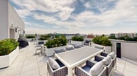 This expansive rooftop terrace is a thoughtfully designed outdoor amenity space that combines contemporary aesthetics with functional gathering areas. The terrace is finished with large-format, light-colored tile pavers that lend a clean, polished look throughout. Multiple distinct zones are arranged across the open deck, each serving a specific purpose while contributing to the overall flow of the space.

A dedicated outdoor dining area features a sleek white rectangular table surrounded by cushioned chairs, ideal for al fresco meals. Nearby, multiple lounge seating arrangements — composed of modern white-framed sofas and chairs with gray upholstered cushions — invite relaxed conversation and leisure. A low-profile fire pit anchors one of the lounge zones, adding ambiance during cooler evenings.

Large white planters filled with lush, manicured shrubs are positioned throughout the terrace, introducing a vibrant layer of greenery that softens the architectural lines and adds visual interest. A gas grill station offers convenient outdoor cooking capability. A glass sliding door provides access to an interior amenity room visible beyond.

The terrace enjoys an open, unobstructed exposure to the sky, with panoramic views of the surrounding urban cityscape stretching across the horizon — a standout feature that elevates this rooftop into a premier outdoor destination.