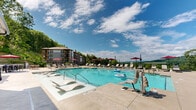 Located at 105 Piney Mountain Dr in Asheville, North Carolina, this outdoor amenity space offers a resort-style experience set against the stunning backdrop of the Blue Ridge Mountains. The centerpiece is a generously sized swimming pool with crystal-clear water, in-pool lounge chairs, and accessibility features including a pool lift, making it welcoming to all residents and guests. The expansive pool deck is lined with cushioned chaise lounges and shaded by red market umbrellas, providing ample space for sunbathing and relaxation. A gated entry with clearly posted pool rules ensures a safe and organized environment. Adjacent to the pool, a covered outdoor lounge area is attached to the building's stone and wood-accented facade, featuring ceiling fans and a wall-mounted television — ideal for casual gatherings in any weather. A dedicated fire pit patio area furnished with plush seating and strung with overhead Edison lights creates an inviting atmosphere for evening socializing, all while overlooking the lush, tree-covered mountain landscape. The surrounding multi-story residential buildings, constructed with a blend of stone masonry and contemporary design elements, frame the space cohesively. The combination of mountain scenery, thoughtful amenities, and well-designed outdoor spaces makes this a standout community destination in the Asheville area.