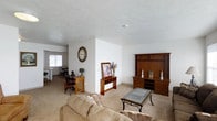 This single-floor space offers a welcoming and functional layout designed for comfortable everyday living. The living area serves as the heart of the home, featuring plush, neutral-toned carpeting that extends throughout and contributes to a warm, cohesive atmosphere. A large wooden entertainment center anchors one wall, providing ample storage and display space, while a glass-top coffee table and a cushioned sofa create an inviting seating arrangement. The space flows naturally into the dining area, which is visible from the living room and benefits from natural light provided by a nearby window. Warm wood furniture pieces, including a side console and decorative accents, complement the room's earthy palette. The textured ceiling adds subtle architectural character throughout. The open-plan design encourages easy movement between the living and dining zones, making the space feel connected and spacious. Neutral white walls keep the interior bright and versatile, allowing the warm wood tones of the furnishings to stand out. Overall, the space presents a tidy, classic residential aesthetic that balances practicality with a relaxed, homey ambiance.