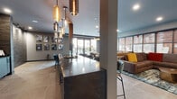 Located at 435 Main St in Hackensack, NJ, this single-floor amenity lounge is a thoughtfully designed communal space that blends contemporary style with inviting warmth. The entrance foyer sets the tone with large-format tile flooring, recessed lighting, and a tasteful potted indoor tree, creating a welcoming first impression.

The main lounge area is anchored by a striking dark marble-veined bar counter, illuminated overhead by an eye-catching cluster of mixed-metal and glass pendant lights in varying heights. This centerpiece island provides flexible seating and serves as a natural gathering hub for the space.

Adjacent to the bar, a generously sized lounge seating zone features plush sectional sofas in leather and upholstered fabrics, accented with colorful throw pillows and layered over richly patterned area rugs. A distinctive feature wall of large-scale paneled imagery adds depth and visual interest to the seating area, complemented by dramatic floor-length navy drapery and a bold gold pendant light fixture above.

Large floor-to-ceiling glass doors flood the interior with natural light and open directly onto an outdoor terrace, where string lights and additional seating create an inviting extension of the lounge. A curated gallery wall of black-and-white framed photographs lines one wall, adding a local artistic flair. The overall ambiance is sophisticated yet comfortable, ideal for socializing or relaxing.