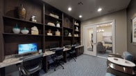 This single-floor business center is thoughtfully designed to support productivity and collaboration in a professional yet welcoming environment. The main workspace is anchored by a built-in dark wood workstation wall, where a granite-topped desk runs the length of the room, accommodating multiple computer terminals paired with ergonomic task chairs. Floor-to-ceiling open shelving above the workstation displays an curated arrangement of books, potted plants, decorative vessels, and sculptural accents, adding warmth and visual interest to the functional setup. Recessed lighting illuminates the space evenly, complemented by natural light filtering through slatted window blinds along the adjacent wall. Soft blue-grey patterned carpet flows throughout, tying the space together cohesively. A set of glass-paneled French doors opens into a private conference room, furnished with a round table and upholstered chairs beneath a chandelier, ideal for small meetings or collaborative sessions. Near the entrance, a cozy lounge nook features light blue accent chairs and a marble-topped side table set against a deep charcoal accent wall adorned with framed artwork. An exterior door provides convenient direct access to the outdoors, completing this well-appointed and versatile business center.