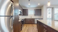This thoughtfully designed single-floor residence showcases a cohesive modern aesthetic throughout its well-proportioned layout. The kitchen is a standout feature, offering rich dark-toned cabinetry with sleek bar-pull hardware, light granite countertops, and a full suite of stainless steel appliances including a refrigerator, range, built-in microwave, and dishwasher. A central island with a matching granite surface provides additional workspace and casual seating potential, while pendant lighting adds a refined touch overhead. Wood-look flooring runs throughout the kitchen and living areas, contributing warmth and visual continuity. Built-in open shelving near the living area offers functional display space. The bedrooms are finished with plush neutral carpeting, ceiling fans, and windows fitted with horizontal blinds that allow natural light while maintaining privacy. The bathrooms mirror the kitchen's design language, featuring dark cabinetry, granite vanity tops, large-format tile surrounds in the shower/tub combination, and wood-look flooring. One bathroom provides direct access to a generously sized walk-in closet, carpeted and equipped with a full-length hanging rod and shelving. The overall palette of whites, warm wood tones, and dark cabinetry creates a clean, contemporary living environment.