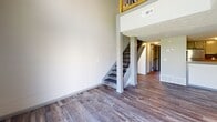 This two-story space is thoughtfully arranged across two levels, with the main living areas on the lower floor and the bedroom suite above. The lower level features an open living area distinguished by soaring ceilings, recessed lighting, and a full-width sliding glass door that floods the space with natural light and provides direct outdoor access. Adjacent to the living area, the galley-style kitchen is well-appointed with granite countertops, a stone tile backsplash, gray cabinetry, stainless steel appliances, and a deep double-basin sink with a matte black faucet. Wood-look flooring runs consistently throughout both floors, adding warmth and visual continuity. A staircase with wood-and-metal railing connects the levels, opening onto an upper-floor hallway with textured ceilings and flush-mount lighting. The generously sized primary bedroom on the upper level features a window with outdoor views, a dedicated closet with built-in shelving and hanging rods, and access to the full bathroom. The bathroom includes a full-size soaking tub with a tiled surround, a vanity with a speckled quartz countertop, an undermount sink, matte black fixtures, and a round mirror, creating a clean, contemporary finish throughout.