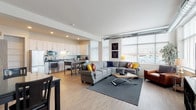 This contemporary two-floor residence offers a thoughtfully designed open-concept layout that seamlessly connects the living, dining, and kitchen areas. The expansive living room is bathed in natural light through large windows with horizontal blinds, and features light hardwood-style flooring, exposed ductwork as an industrial design accent, and ample space for comfortable furnishings. Large corner windows provide broad exterior views and contribute to the bright, airy atmosphere.

The kitchen is well-appointed with white shaker cabinetry, gray countertops, a full suite of stainless steel appliances including a refrigerator, range, microwave, and dishwasher, complemented by a tile backsplash and a convenient kitchen island with seating. Open shelving and generous cabinet storage add practicality to the space.

The bedrooms offer a calm retreat, finished with plush carpeting, natural light from blinds-fitted windows, and closet access. The primary bedroom is particularly serene and spacious.

Two bathrooms are present, both featuring white vanities with solid countertops, wood-look tile flooring, and clean neutral tones. One bathroom includes a shower-tub combination, while the other provides a more streamlined layout. Throughout the unit, a consistent modern aesthetic is maintained with neutral walls and quality finishes.