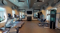 This fitness center is a thoughtfully designed, single-floor facility built to support a comprehensive workout experience. The space features durable speckled rubber flooring throughout, providing both comfort and safety for a wide range of exercises. Overhead, dual ceiling fans and recessed lighting work in tandem to maintain a comfortable environment during intense workouts, while an exposed duct adds an industrial aesthetic touch to the ceiling.

Natural light filters in through multiple windows positioned along the walls, creating a bright and energizing atmosphere. Large, dark-framed mirrors are strategically placed to visually expand the space and allow users to monitor their form during exercise.

The cardio section is well-stocked with multiple treadmills and stationary bikes, each equipped with individual digital displays, as well as elliptical trainers suitable for low-impact workouts. A dedicated strength training area includes a multi-tiered dumbbell rack with a full range of free weights, weight benches, a cable machine, and additional resistance equipment. Stability balls and other accessories are neatly stored on dedicated racks. A wall-mounted flat-screen television provides entertainment for users. The facility is accessed through elegant black-framed double glass doors, reinforcing the polished, modern aesthetic of the overall space.