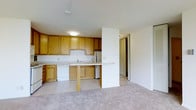 This single-floor residence offers a functional and welcoming layout centered around an open-concept living and kitchen area. The kitchen is well-appointed with warm honey-toned wood cabinetry providing ample storage, a white dishwasher, refrigerator, and a double sink set beneath the upper cabinets. A peninsula breakfast bar extends from the kitchen, creating a natural division between the tiled kitchen floor and the carpeted living area beyond, while maintaining an open and connected feel throughout the space. The light-colored tile in the kitchen transitions smoothly into plush, neutral-toned carpeting in the living area, contributing to a comfortable and cohesive atmosphere. The white walls and ceilings keep the space feeling bright and clean, with flush-mounted ceiling lighting providing consistent illumination. A doorway visible from the main living area leads further into the residence, suggesting additional private rooms beyond. The overall design is practical and straightforward, offering a layout that balances everyday functionality with a comfortable living environment suited for a variety of lifestyles.